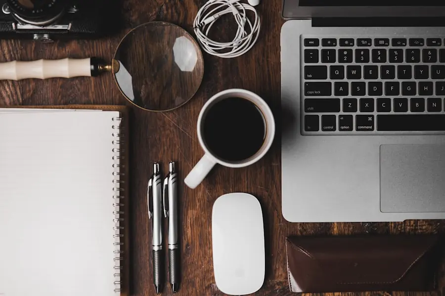 A paper notebook, pens and a magnifying glass laid out beside a laptop on a desk