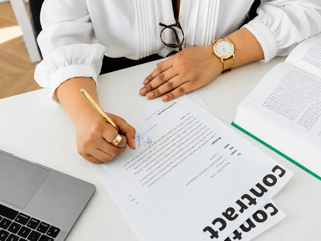 A contract being signed at a desk beside a laptop